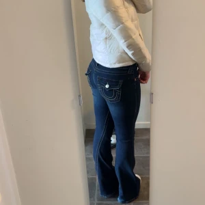 True Religion Joey Low Rise Flare Jeans - Mörkblå jeans från True Religion, modell Joey med låg midja och utsvängda ben. Snygga kontrastsömmar och ikoniska fickdetaljer med knappar bak. Klassisk femficksmodell. Storlek 26. Lite trampade på men det är inget som förstör byxorna! 💗💗💗 Säljer då de är för stora på mig 