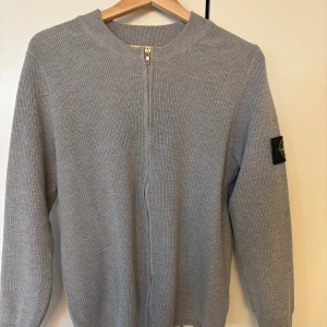 Stone island kofta  - Snygg grå stickad kofta från Stone Island med dragkedja framtill och klassisk patch på ärmen. Perfekt för lager-på-lager och enkel att matcha med olika outfits. Ribbade muddar och rund halsringning.