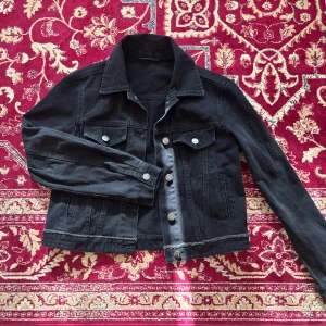 Svart jeansjacka från MO&CO - Säljer en svart jeansjacka från MO&CO med klassisk krage, silverfärgade knappar och två bröstfickor med lock. Jackan har en rak, något croppad passform och långa ärmar. Perfekt att slänga på sig över en hoodie eller t-shirt.