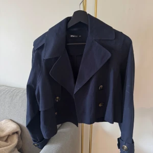 Mörkblå croppad trenchcoat från Gina Tricot - Säljer en mörkblå croppad trenchcoat från Gina Tricot. Använd Max 3 gånger men kommer aldrig till användning längre! Jackan har dubbelknäppning med stora knappar, klassisk krage och långa ärmar med spänne vid ärmslut. Perfekt för vår och höst.