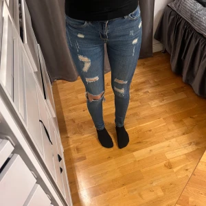 Blå slitna skinny jeans - Säljer ett par blå lågmidjade skinny jeans med slitningar och hål på knäna. Jeansen har normal passform i midjan och är i klassisk denim med dragkedja och knapp framtill (tappat lite färg). Storlek Xs från Lager157.