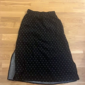 Svart prickig långkjol från 157 - Svart långkjol från 157 med vita prickar och resår i midjan. Kjolen har en slits på sidan som ger en snygg detalj. Perfekt för dig som gillar klassiska mönster och enkel stil.