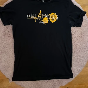 Svart t-shirt med tryck från Jack & Jones - Svart t-shirt från Jack & Jones Originals med vit och gul tryckt text och rosdetalj framtill. Klassisk passform och korta ärmar. Perfekt för en stilren och avslappnad look.