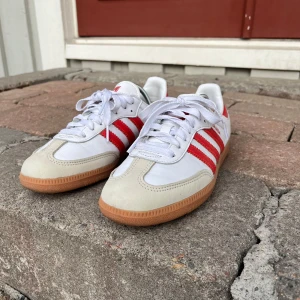 Adidas Samba OG W - låga sneakers med röda ränder - Säljer ett par Adidas Samba sneakers i vitt skinn med röda detaljer och ränder. Skorna har den klassiska gummisulans design, snörning och beige mockadetaljer vid tån, vilket ger dem en tidlös retrolook. De är i mycket bra skick, nästan som nya, och har bara använts ett fåtal gånger. Storleken är 39,5. Ett perfekt val för dig som vill ha ett par stiliga och sportiga sneakers med både komfort och klass. Passar lika bra till vardags som till mer avslappnade tillfällen.