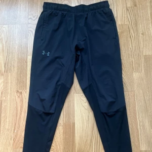 Svarta träningsbyxor från Under Armour - Säljer ett par svarta träningsbyxor från Under Armour med elastisk midja och smal passform. Byxorna har diskret logga på låret och paneler i olika material för extra rörlighet. Perfekta för gym eller löpning.