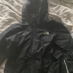Svart vindjacka från The North Face - Säljer en svart vindjacka från The North Face med huva och dragkedja framtill. Jackan har två sidofickor med dragkedja och logga på bröstet. Perfekt för blåsiga dagar!
