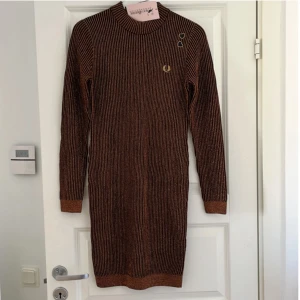 Randig stickad klänning från Fred Perry x Amy Winehouse Foundation - Säljer en stickad långärmad klänning från Fred Perry i samarbete med Amy Winehouse Foundation. Klänningen är svart med kopparfärgade ränder, har ribbad struktur och rund hals. Broderad logga på bröstet och två hjärtformade detaljer vid axeln.