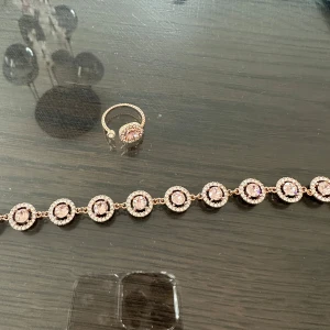 Glittrigt armband och ring i roséguld - Säljer ett matchande set med armband och ring i roséguld. Båda har runda, ljusrosa stenar i mitten omgivna av små gnistrande stenar för extra bling. Perfekt för dig som gillar eleganta och glittriga accessoarer med lyxig känsla.