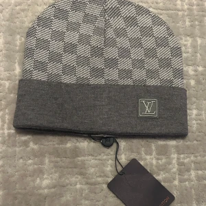 Grå rutig mössa från Louis Vuitton - Exklusiv grå och vit rutig mössa från Louis Vuitton med LV-logga framtill. Mössan har bred uppvikt kant och är stickad i mjukt material som känns lyxigt. Perfekt accessoar för att lyfta din vinterstil.