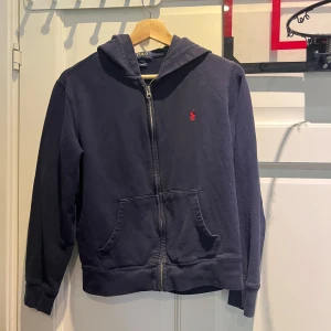 Mörkblå hoodie från Polo Ralph Lauren - Mörkblå hoodie från Polo Ralph Lauren med dragkedja framtill och klassisk röd logga broderad på bröstet. Tröjan har huva, känguruficka och är tillverkad i mjuk bomull. Perfekt för en chill och stilren look.
