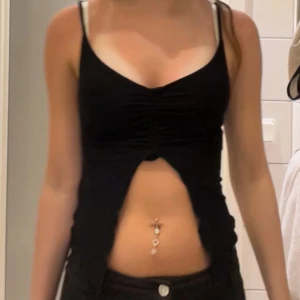Topp - Säljer en svart croppad linnetopp med smala axelband och snygg snörningsdetalj framtill. Toppen har en djup v-ringning och öppen front som ger en edgy look. Perfekt för dig som gillar trendiga och unika plagg.