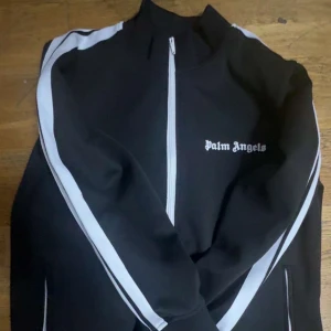Palm Angels svart track jacket - Svart track jacket från Palm Angels med vita ränder längs ärmarna och vit dragkedja. Jackan har hög krage, två fickor med dragkedja och logga på bröstet. Perfekt för en sportig och clean streetstyle. Fick den i present men den passade inte så jag kommer se till och sälja den, inga skador helt ny använt den 1-2 gånger men den passar inte! Pris kan diskuteras vid snabb affär