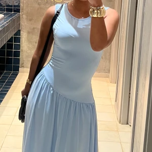 Ljusblå långklänning med asymmetrisk midja - Supersöt ljusblå långklänning med ärmlös design och rund halsringning. Klänningen har en snygg asymmetrisk midja och ett mjukt, flowy fall. Perfekt för dig som gillar en clean och fräsch look med en twist.
