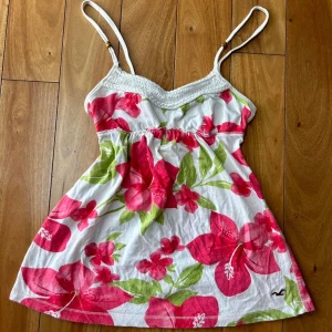 Blommig babydoll från Hollister - Supersöt linnetopp från Hollister med stora rosa och röda blommor på vit botten. Smala axelband och lätt rynkad upptill, perfekt för varma dagar. Liten broderad logga nertill. Lätt och luftig känsla med somriga vibes. Inga bilder på!!