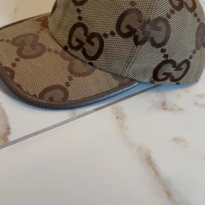 Gucci keps med GG-monogram - Exklusiv keps från Gucci i beige och brunt med det klassiska GG-monogrammet över hela kepsen. Tillverkad i bomullscanvas med bruna detaljer och böjd skärm. Kommer med originalpåse och tags. Perfekt för dig som vill sticka ut med lyxig streetstyle.