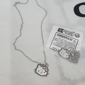 Hello Kitty halsband med strass - Snyggt halsband med Hello Kitty-hänge täckt av glittrande strass. Hänget har en söt rosa rosett och detaljer i svart och gult. Kedjan är i silverfärgad metall och passar perfekt för dig som gillar kawaii-stil och vill sticka ut lite extra.
