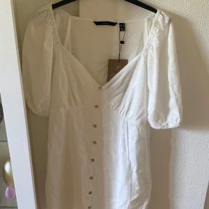 Vit kortklänning med puffärm - Superfin vit kortklänning från Lindex med puffärmar, v-ringning och knappar framtill. Klänningen har en luftig känsla och är perfekt för varma dagar. Tillverkad i bomull och har en feminin siluett med fina detaljer.