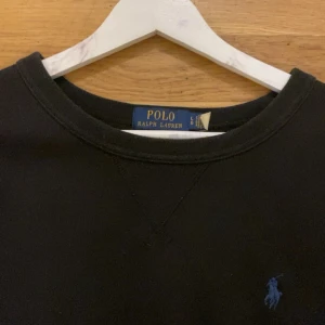 Svart sweatshirt från Polo Ralph Lauren - Klassisk svart sweatshirt från Polo Ralph Lauren med rund halsringning och diskret blå logga broderad på bröstet. Tröjan har långa ärmar och en relaxed passform, perfekt för en stilren och enkel look. Tillverkad i mjuk bomull för maximal komfort.