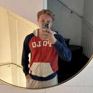 Röd och blå hoodie från Polo Ralph Lauren - Säljer en hoodie från Polo Ralph Lauren i rött och marinblått med vit huva och ficka. Stor POLO-text framtill, dragkedja och känguruficka. Perfekt för dig som gillar sportig och avslappnad stil.