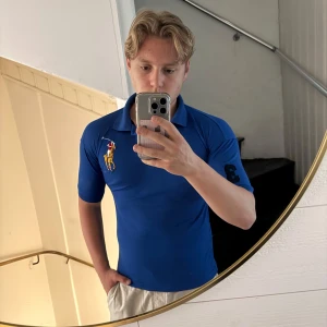 Blå pikétröja från Polo Ralph Lauren - Snygg blå pikétröja från Polo Ralph Lauren med klassisk krage och korta ärmar. Stor broderad polospelare på bröstet och broderad siffra på ärmen. Materialet är bomull och tröjan har en normal passform. Perfekt för en sportig och stilren look. Sitter som XS/S