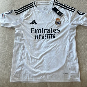 Real Madrid Mbappé 9 matchtröja Adidas - Säljer en officiell Real Madrid fotbollströja från Adidas med Mbappé och nummer 9 på ryggen. Tröjan är vit med svarta detaljer, klubbmärke, Champions League-märke och sponsortryck. Tillverkad i lätt och ventilerande material med korta ärmar och klassiska Adidas-ränder på axlarna.