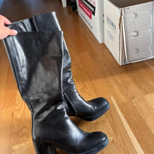 Svarta höga stövlar med klack - Snygga svarta stövlar i skinn med hög klack och rund tå. Stövlarna har dragkedja på insidan och en stilren, klassisk look som passar till många outfits. Perfekta för dig som vill ha en tidlös och edgy stil.