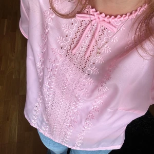 Rosa blus - Supersöt ljusrosa blus i satinliknande material med broderade detaljer framtill, liten rosett vid halsen och knappar i ryggen. Blusen har korta ärmar. 