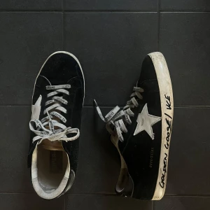 Golden Goose  - Svarta sneakers från Golden Goose med vit stjärndetalj på sidan och text på sulan. Skorna har grå snörning med logga och en vit platt sula. Ovandelen är i mocka och insidan är ljus. Perfekta för dig som gillar streetstyle och unika detaljer.