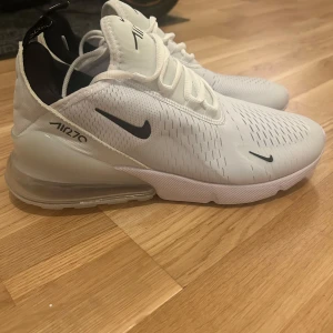 Nike Air Max 270 vita sneakers - hellt nya Nike Air Max 270 sneakers i vitt med svarta detaljer och stor synlig Air-enhet i hälen. Ovandel i mesh för bra andningsförmåga, snörning och modern design. Perfekta för dig som gillar stilrena och sportiga skor med maximal komfort.
