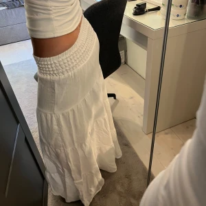 Vit långkjol från H&M med smock - Superskön vit långkjol från H&M med bred smock i midjan och lätt, flowigt tyg. Kjolen är i flera lager och har en luftig känsla, perfekt för varma dagar. Materialet är en mix av viskos och polyamid som ger ett mjukt fall.