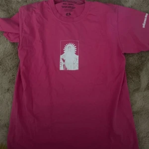 Rosa t-shirt med tryck från Yung Lean  - Rosa t-shirt från Unhuman med vit grafisk print av en figur med gloria på bröstet. Klassisk passform och rund halsringning. Tryck även på ena ärmen. Perfekt för dig som gillar streetwear och unika prints.