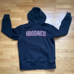 Svart HOODRICH hoodie med tryck - Svart hoodie från HOODRICH i storlek S med stor vit och röd logga på bröstet. Klassisk känguruficka framtill, huva med snörning och långa ärmar. Perfekt för dig som gillar streetwear och vill sticka ut med en bold look.