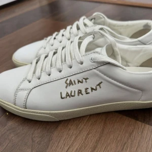 Vita Saint Laurent sneakers i skinn - Snygga vita sneakers från Saint Laurent med handskriven logga på sidan. Skorna har klassisk låg profil, rund tå och vita snören. Tillverkade i mjukt skinn med en stilren och minimalistisk look som passar till det mesta.