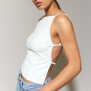 Vit  topp med öppen rygg - Säljer en vit croppad linnetopp med smala axelband och öppen rygg. Toppen har minimalistisk design med justerbara band i ryggen och sidorna, vilket ger en snygg och modern look. Perfekt till jeans eller kjol för en trendig stil. helt oanvänd 