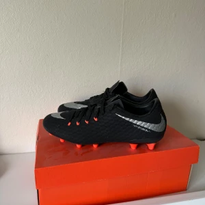 Nike Hypervenom svart fotbollssko - Svarta Nike Hypervenom fotbollsskor med silvriga swoosh-detaljer och orange dobbar. Ovandelen är i syntetmaterial med textur för bättre bollkontroll och snörning framtill. Snygg och sportig design som sticker ut på planen.