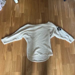 Beige stickad tröja med stjärndetalj - Säljer en beige stickad tröja i 100% bomull. Tröjan har lång ärm och en cool stjärnpatch på ena ärmen. Modellen är loose fit med rundad nederkant och bred halsringning. Perfekt för dig som gillar en avslappnad och trendig stil.