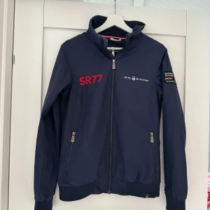 Marinblå vindjacka Sail Racing SR77 - Snygg marinblå vindjacka från Sail Racing med SR77-tryck och broderade detaljer. Jackan har dragkedja, ribbade muddar och två fickor med dragkedja. Fodret är rött med Sail Racing-tryck. Perfekt för dig som gillar sportig och stilren look. Använd fåtal gånger. Köpt för 2400kr