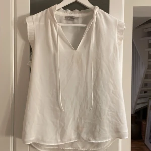 TRYCK EJ PÅ KÖP! Skriv och köp varan hos Alicen!  - Vit ärmlös blus från H&M med plisserad krage och ärmslut. Blusen har v-ringning med tunna knytband framtill och är tillverkad i ett lätt, flowy material som ger en luftig känsla. Perfekt för en clean och stilren look.