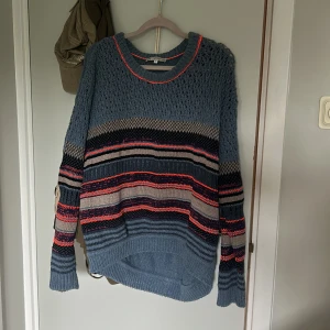 Randig stickad tröja från Dagmar XS - Säljer en oversized stickad tröja från Dagmar i blått med randigt mönster i blått, rosa och grått. Tröjan har lång ärm, rund hals och coola virkade detaljer upptill. Perfekt för dig som gillar unika och färgglada plagg.