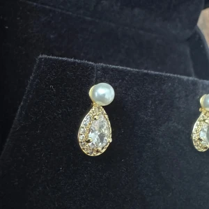 Guldiga örhängen med pärla och kristall - Ett par vackra örhängen med en droppformad design. Örhängena är prydda med gnistrande kristaller och en elegant pärla på toppen.   925 silver pläterad med gult guld plätering samt zirconia kristaller därmed en icke allergisk pärla.   Shell Bredd 4mm  Örhängen 12,5mm*6,5mm