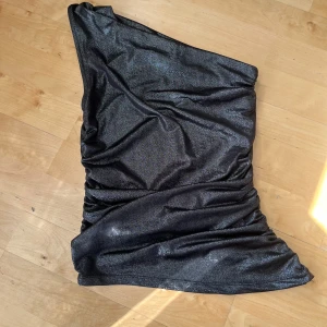 Glittrig svart oneshoulder topp - Säljer en glittrig svart oneshoulder topp med tight passform och snygga rynkade detaljer längs sidorna. Toppens material har en skimrande finish som fångar ljuset och ger en festlig vibe. Perfekt för dig som vill sticka ut med en trendig och cool look.