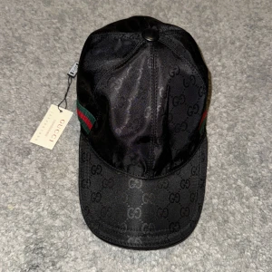 Svart keps från Gucci med GG-mönster - Snygg svart keps från Gucci i glansigt material med klassiskt GG-monogram över hela kepsen. Har gröna och röda ränder på sidan och justerbart spänne bak. Perfekt för dig som vill ha en exklusiv och trendig accessoar.