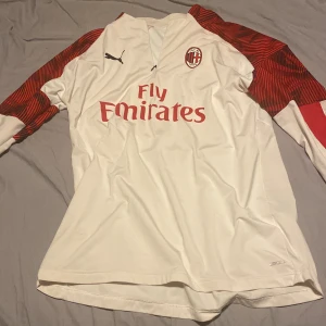 AC Milan långärmad bortatröja Puma - Hel äkta AC Milan långärmad bortatröja från Puma i vitt med röda och svarta grafiska detaljer på ärmarna. Tröjan har klubbmärke på bröstet, Fly Emirates-tryck och dragkedja vid halsen. Tillverkad i lätt och ventilerande funktionsmaterial.