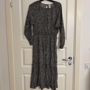 Klänning - Midiklänning från H&M i svartvitt mönster med små prickar. Klänningen har lång ärm, rund halsringning och knäppning bak i nacken. Resår i midjan ger en snygg siluett och tyget är lätt och luftigt, perfekt för vår och sommar.