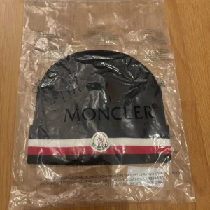 Svart Moncler mössa  - Svart Moncler mössa med klassiska ränder i marinblått, vitt och rött längs kanten. Mössan har Moncler-logga framtill och är ribbstickad för en snygg passform. Perfekt accessoar för att lyfta din vinterstil.