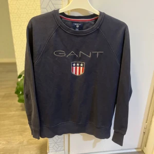 Mörkblå sweatshirt från GANT - Snygg mörkblå sweatshirt från GANT med broderad logga och sköld på bröstet. Tröjan har rund halsringning, långa ärmar och klassisk passform. Perfekt för en chill och stilren look. Materialet är mjuk bomull som känns skönt mot huden.