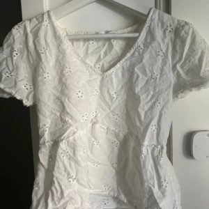 Vit blus med broderade blommor - Söt vit blus med v-ringning och korta ärmar. Blusen har broderade blommor över hela tyget och en lätt volang vid midjan. Perfekt för en somrig och fräsch look.