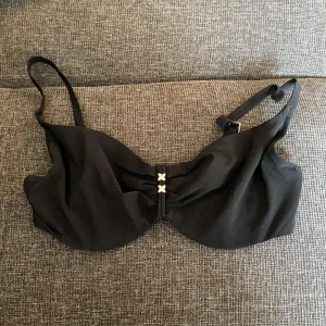 Svart bikiniöverdel med detaljer - Svart bikiniöverdel med justerbara axelband och snygga små kryssdetaljer framtill. Klassisk form med bygel och knäppning bak. Perfekt för stranden eller poolen, enkel att matcha med olika underdelar. Kan mötas upp i Trollhättan eller Alingsås. Kan skickas men då tillkommer en fraktkostnad.