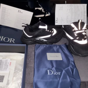 Svarta sneakers från Dior med reflexdetaljer - Chunky svarta sneakers från Dior med coola reflekterande detaljer och meshpaneler. Skorna har snörning, rund tå och en kraftig platt sula. Snygg kontrast mellan svart och vitt, perfekt för dig som gillar statement-sneakers med lyxig känsla.