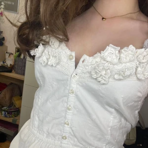 Populär hollister topp med blommor  - Superfin vit linnetopp med broderade blommor och spetsdetaljer vid urringningen. Linnet har knappar framtill, justerbara axelband och en lätt rynkad midja för en söt siluett.  Linnet är ifrån hollister och säljs inte mer ❤️